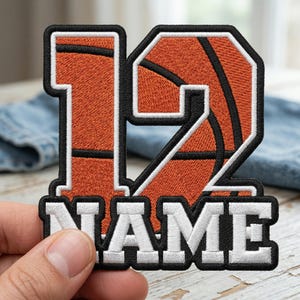 May include: Embroidered patch featuring the number 12 in orange, with a basketball design. The number is outlined in black and white. Below the number is the word "NAME" in white block letters. The patch is likely for sports apparel.