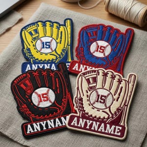 May include: Four baseball glove patches in yellow, blue, black, and cream colors. Each patch features a baseball with the number 15 and a banner with text. The patches are on a gray fabric surface.