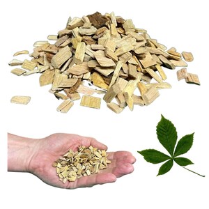 Puede incluir: Un montón de virutas de madera de color claro, con algunas piezas más oscuras, adecuadas para ahumar o asar a la parrilla. Una mano sostiene una pequeña cantidad de virutas, y también hay una hoja verde.