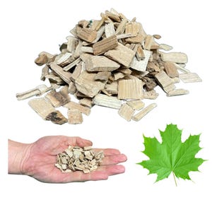 Puede incluir: Un montón de virutas de madera de color marrón claro, con un puñado en la mano de una persona y una hoja de arce verde. Las virutas de madera son probablemente para ahumar o asar a la parrilla.
