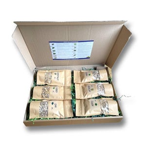 Puede incluir: Una caja de cartón abierta llena de varias bolsas de papel marrón selladas con virutas de madera para ahumar. Las bolsas están etiquetadas con diferentes tipos de madera: Castaño, Cerezo, Roble, Nogal americano, Manzano y Arce.