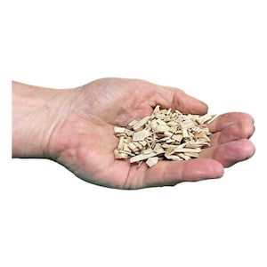 Puede incluir: Primer plano de una mano sosteniendo un puñado de virutas de madera claras. Las virutas, de formas y tamaños variados, tienen un aspecto natural y sin tratar. El fondo es blanco liso.