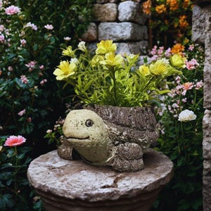 Könnte beinhalten: Ein steinerner, schildkrötenförmiger Blumentopf, gefüllt mit gelben Blumen und grünen Blättern. Der Topf ist hellgrün und braun, mit einem strukturierten Panzer. Er steht auf einem Steinsockel in einem Garten.