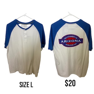 Puede incluir: Camiseta de estilo raglán blanca y azul. La camiseta tiene mangas azules y un cuerpo blanco con cuello Henley. La parte trasera de la camiseta presenta un logotipo de la Universidad de Arizona. La camiseta es talla L.