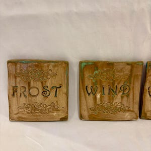 May include: Square ceramic tiles with the words "FROST" and "WIND" in a rustic font. The tiles are brown with a green border and floral designs. The tiles are likely decorative elements.