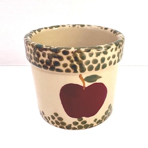 Può includere: Vaso di fiori in ceramica color crema con un grande motivo a mela rossa. Il bordo e la base sono decorati con pois verdi e marroni. La mela ha una foglia verde e un gambo marrone.