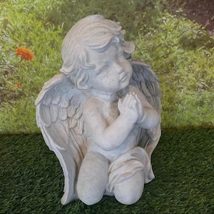 May include: A kneeling cherub statue with folded hands, wings, and a serene expression. The statue is made of a light gray material and is positioned on a bed of green grass, with a blurred background of greenery and a hint of an orange flower.