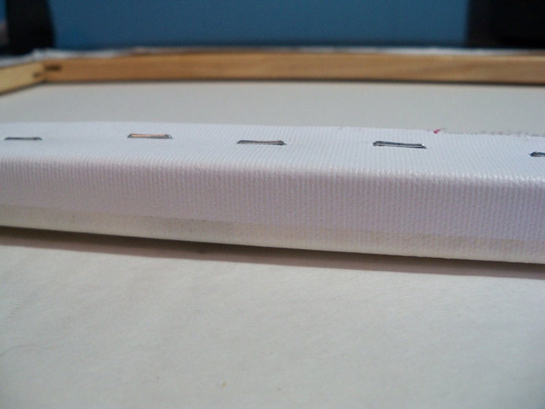 May include: Close-up of a white canvas stretched over a wooden frame. The canvas is secured with staples along the edges.