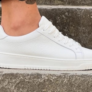 Może przedstawiać: Biały skórzany sneaker z teksturowaną cholewką, grubą podeszwą i białymi sznurowadłami. But jest czysty i wygląda na nowy, o minimalistycznym designie. Sneaker to opcja obuwia na co dzień.