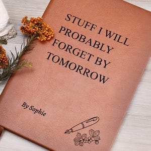 May include: A tan leather journal with the text "STUFF I WILL PROBABLY FORGET BY TOMORROW" in black. The journal also has the name "By Sophie" and a pen and floral illustration. A brown ribbon bookmark is visible.