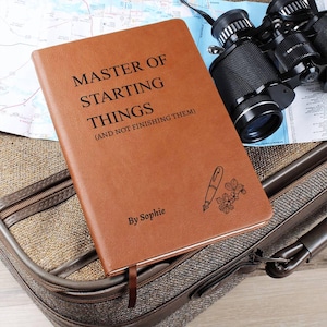 May include: A tan leather journal with the text "MASTER OF STARTING THINGS (AND NOT FINISHING THEM)". The journal features a pen and flower illustration and a ribbon bookmark. The journal is on a suitcase.