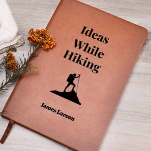 May include: A brown leather journal featuring the words "Ideas While Hiking" and a silhouette of a hiker on a mountain. The name "James Larson" is printed below. A brown ribbon bookmark is visible.