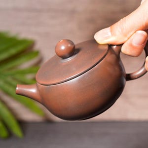 May include: A small, brown ceramic teapot with a rounded body, a curved spout, and a loop handle. The lid has a small, round knob. The teapot is held by a hand, suggesting its size and use for tea.
