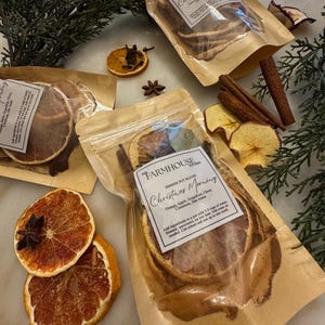 Puede incluir: Bolsas transparentes llenas de rodajas de naranja secas, palitos de canela y anís estrellado. Las bolsas están etiquetadas como "Christmas Morning" y "Simmer Pot Blend". Ramas de hoja perenne y rodajas de manzana secas decoran la escena.