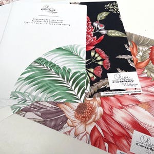 May include: A collection of wallpaper samples featuring various botanical prints. One sample displays green palm leaves on white, while others show red and orange protea flowers on black and white backgrounds. Each sample includes the Olive and the Cowboy logo.