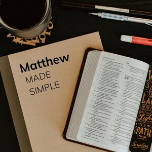 May include: A tan book with the words "Matthew MADE SIMPLE" in black text is next to an open Bible. A cup of coffee and pens are in the background. The Bible has a brown leather cover and a bookmark.