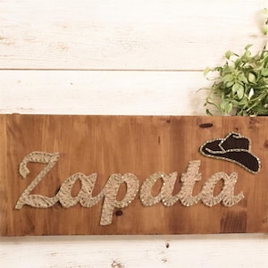 May include: Wooden sign with the name "Zapata" crafted from twine, featuring a cowboy hat graphic. The sign is mounted on a stained wooden board, with a rustic aesthetic. The background is a white wooden surface.