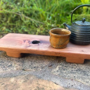 May include: A wooden tea tray with a small, round hole. A small, textured, ceramic tea cup and a black cast iron teapot with a handle sit on the tray. The tray is made of light brown wood.