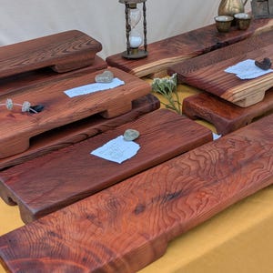 May include: A display of handcrafted wooden serving boards with a rich, reddish-brown hue. The boards vary in size and shape, showcasing the natural wood grain. Some boards have small, decorative stones and paper tags. The boards are displayed on a yellow surface.