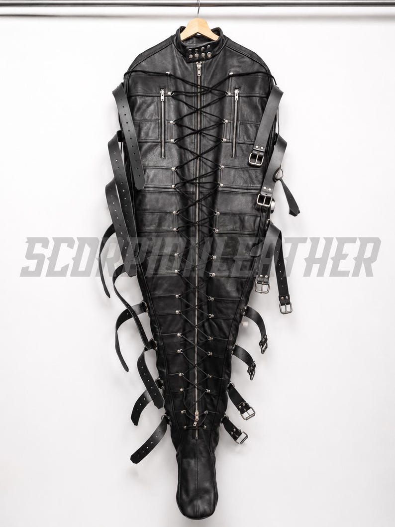 May include: A black leather full-body restraint suit featuring a central vertical zipper, extensive black lace-up detailing, multiple adjustable buckled straps along the sides, and two zippered chest pockets. The text SCORPION LEATHER is visible.