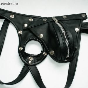 May include: Black leather jockstrap with a zippered pouch and silver-tone metal hardware. The jockstrap features a circular opening and adjustable straps. The text "og scorpionleather" is visible in the upper left corner.
