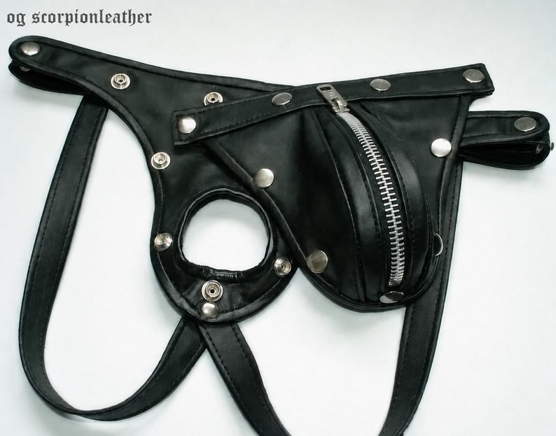 May include: Black leather jockstrap with a zippered pouch and silver-tone metal hardware. The jockstrap features a circular opening and adjustable straps. The text "og scorpionleather" is visible in the upper left corner.
