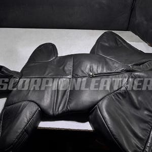 May include: A black leather garment featuring a hood, long sleeves, and a central zipper closure. The piece is constructed with multiple stitched panels and includes the text SCORPIONLEATHER printed across the chest area in a bold, sans-serif font.