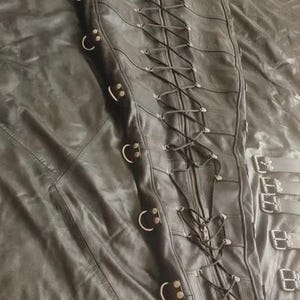 May include: A black leather garment featuring a central vertical zipper and a long, crisscrossing lace-up closure system. The sides are adorned with multiple metal D-rings, and the piece includes additional buckled straps along the side edge.