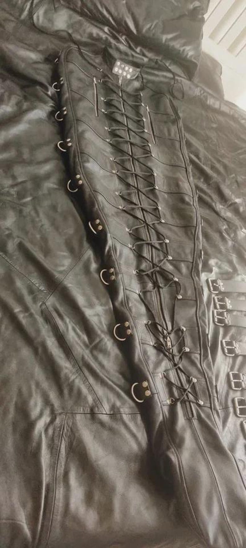 May include: A black leather garment featuring a central vertical zipper and a long, crisscrossing lace-up closure system. The sides are adorned with multiple metal D-rings, and the piece includes additional buckled straps along the side edge.