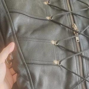 May include: A black leather garment featuring a central metal zipper closure flanked by a decorative crisscross lacing system secured with silver-toned metal eyelets. The piece includes horizontal stitched panels and side D-rings for attachment.