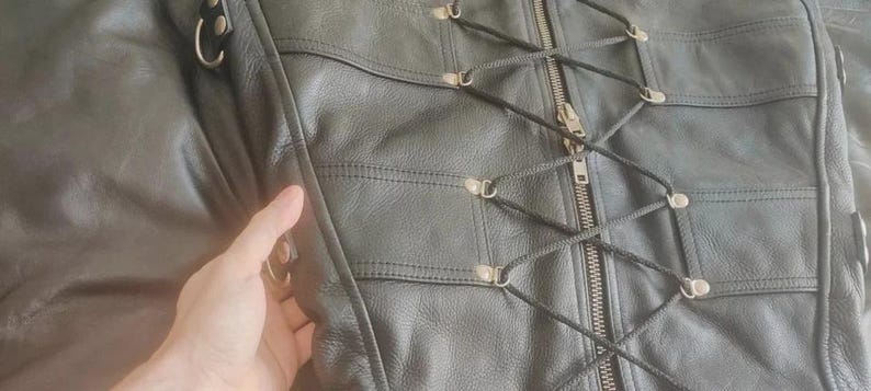 May include: A black leather garment featuring a central metal zipper closure flanked by a decorative crisscross lacing system secured with silver-toned metal eyelets. The piece includes horizontal stitched panels and side D-rings for attachment.