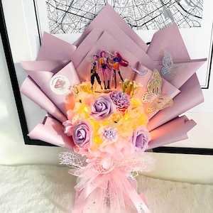 May include: A decorative bouquet of light pink and purple faux flowers, wrapped in light pink paper. The bouquet is decorated with figurines, butterflies, and a ribbon that says "Sweet!"