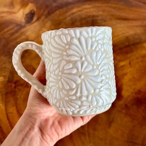 Puede incluir: Una taza de cerámica con una base de color crema brillante y un patrón floral de pétalos blancos en relieve que cubre la superficie exterior. La taza incluye un asa curva con un diseño de puntos texturizados a juego en el borde.