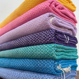 May include: A stack of seven folded textile cloths featuring a consistent diamond weave pattern. The colors from top to bottom are mustard yellow, bright pink, light lavender, deep purple, teal, royal blue, and light periwinkle blue.