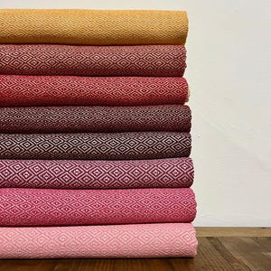 May include: A vertical stack of eight folded textiles, each featuring a consistent white diamond geometric pattern. The colors transition from a golden yellow at the top, through various shades of red and burgundy, to pink at the bottom.