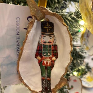 Puede incluir: Adorno navideño hecho a mano con un diseño de soldado cascanueces dentro de una concha de ostra blanca con ribete dorado. El adorno cuelga de una cinta dorada y tiene un diseño festivo. El texto "CUSTOM HANDMADE" es visible.