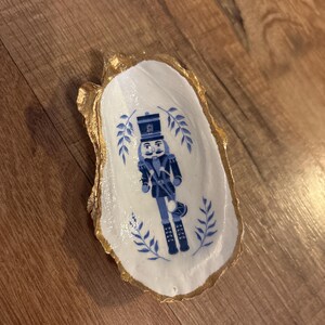 Pode incluir: Uma concha de ostra decorativa com borda pintada de ouro. O interior branco apresenta uma ilustração azul e branca de um soldado quebra-nozes e detalhes de folhas. Uma tigela de bugigangas artesanal única.
