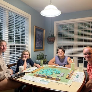 Könnte beinhalten: Vier Personen spielen Mahjong an einem Tisch. Das Spielbrett ist grün mit blauen Steinen. Der Raum ist hell und einladend, mit natürlichem Licht, das durch die Fenster scheint.