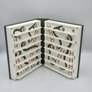 May include: A unique, open book-shaped game with a green cover and white interior. The interior features a series of shelves with green snake-like designs, white circular holes, and the letter "H" in brown. A star is visible on the right side.