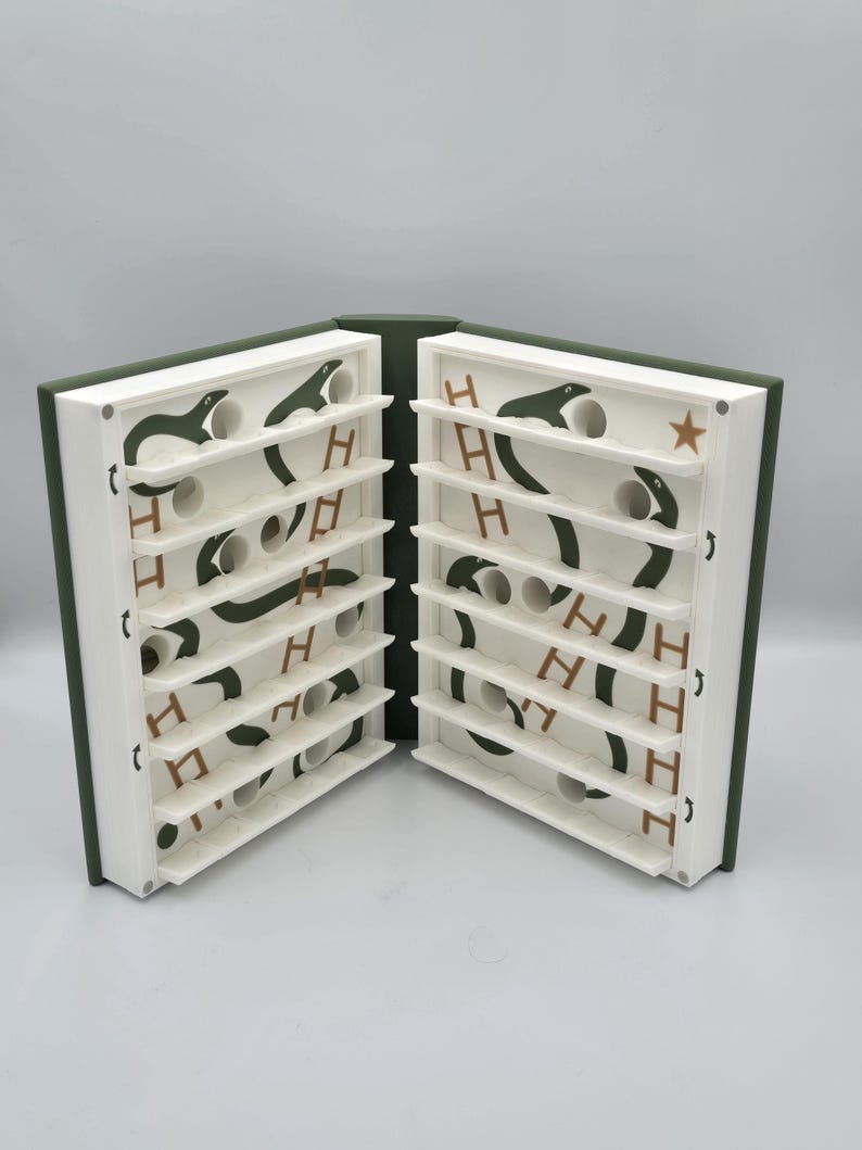 May include: A unique, open book-shaped game with a green cover and white interior. The interior features a series of shelves with green snake-like designs, white circular holes, and the letter "H" in brown. A star is visible on the right side.