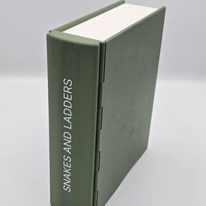 May include: A closed, olive green book with the title "Snakes and Ladders" printed vertically on the spine. The book has a white page edge and a hinged spine.