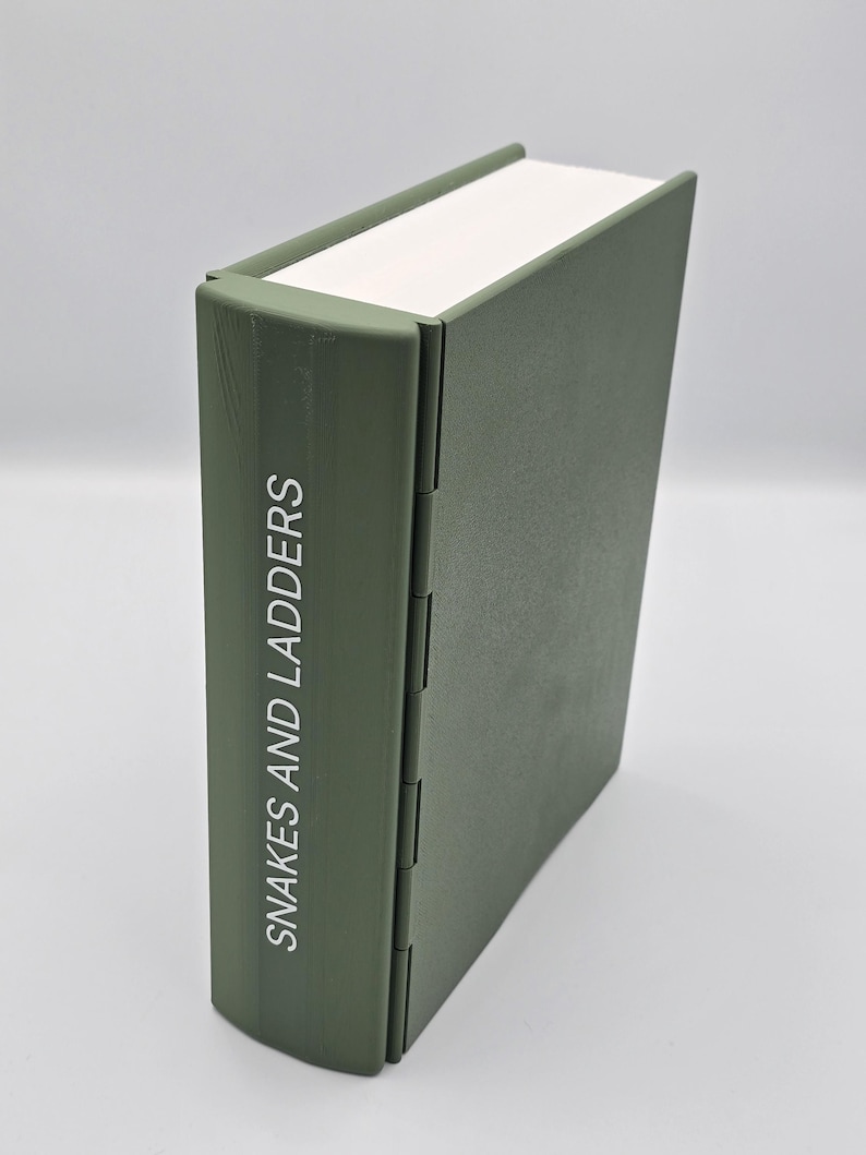 May include: A closed, olive green book with the title "Snakes and Ladders" printed vertically on the spine. The book has a white page edge and a hinged spine.
