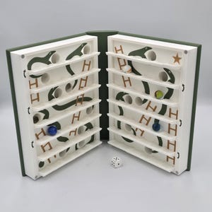 May include: A wooden marble game with a book-like design, open to reveal two white game boards. Each board features a green snake design, shelves, and holes for marbles. A die sits between the boards, and marbles of various colors are visible.