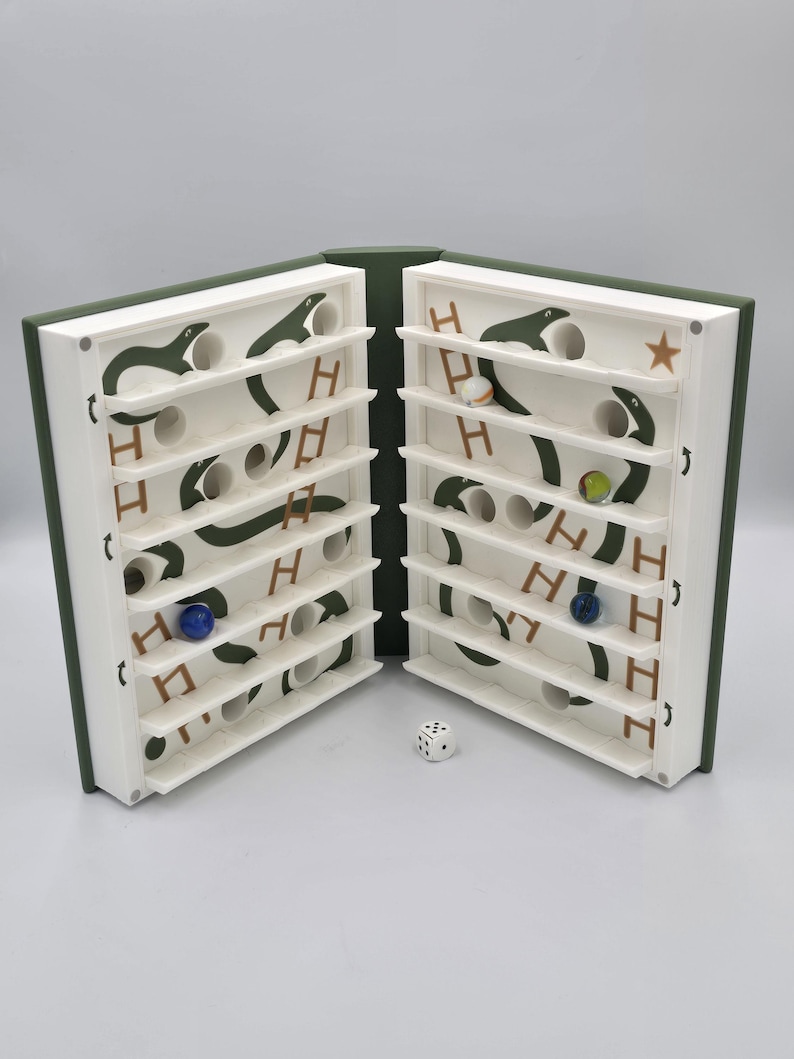 May include: A wooden marble game with a book-like design, open to reveal two white game boards. Each board features a green snake design, shelves, and holes for marbles. A die sits between the boards, and marbles of various colors are visible.