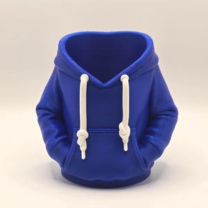 May include: A cobalt blue hoodie-shaped planter with white drawstrings and a front pocket. The miniature hoodie has a hood and is designed to hold small plants or succulents. The planter is a unique decorative item.