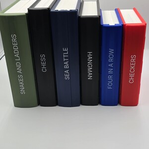 May include: A collection of six board game books with white pages and colorful spines. The titles include "Snakes and Ladders", "Chess", "Sea Battle", "Hangman", "Four in a Row", and "Checkers". The spines are in various colors, including green, black, blue, and red.
