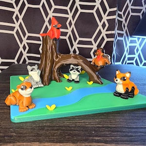 May include: A colorful diorama featuring woodland creatures. A red cardinal sits atop a brown tree stump. Other animals include a fox, raccoons, and an otter, all arranged on a green base with a blue river and yellow leaf accents.