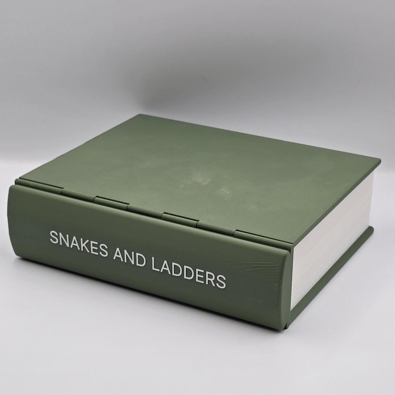 May include: A decorative box designed to look like a book. The box is olive green with the words "SNAKES AND LADDERS" in white lettering on the spine. The pages are white, and the box is closed.