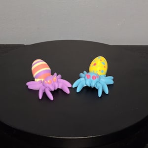 以下が含まれることがあります： 2つのカラフルな粘土製のクモの置物。1つは紫色の体と脚、オレンジと黄色のストライプの卵型の頭を持っています。もう1つは青い体と脚、青い水玉模様の黄色の卵型の頭を持っています。