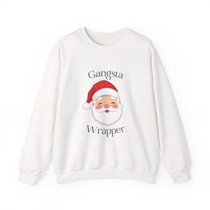 Könnte beinhalten: Weißes Sweatshirt mit einer Grafik von Santa Claus mit roter Mütze und weißem Bart. Der Text "Gangsta Wrapper" befindet sich über und unter der Santa-Grafik. Das Sweatshirt hat lange Ärmel und einen Rundhalsausschnitt.